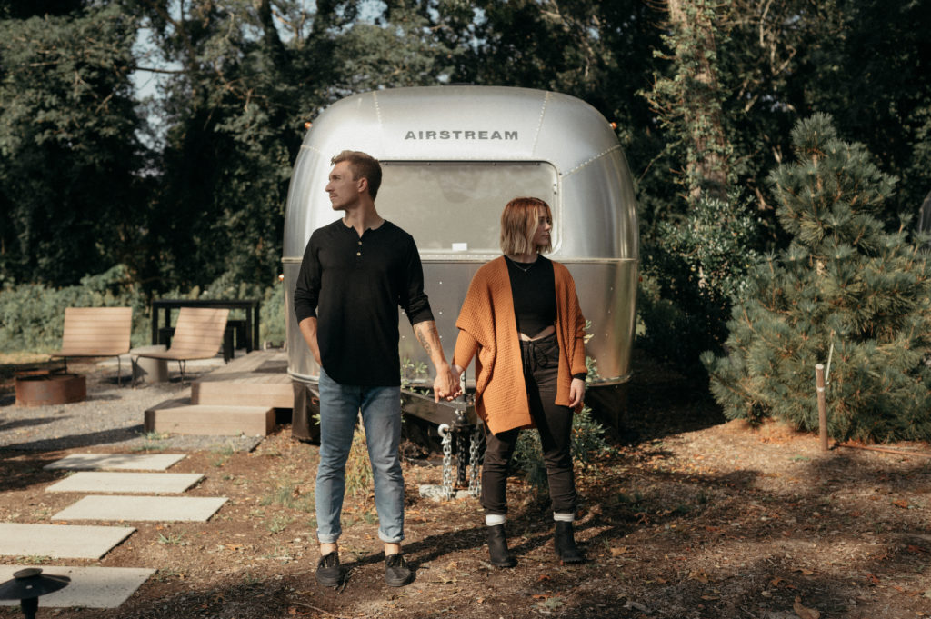 man and woman posing in front of an airstream holding hands. man in black shirt and blue jeans and woman in all black and orange cardigan.