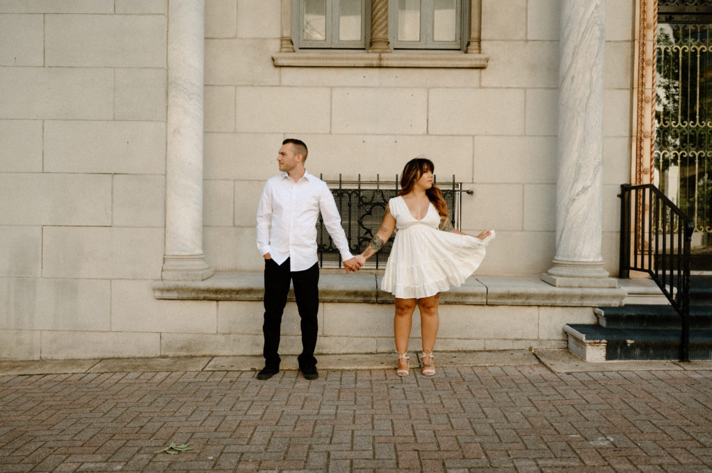 man and woman posing in the city. man in black jeans and white shirt, woman in white flowy dress 