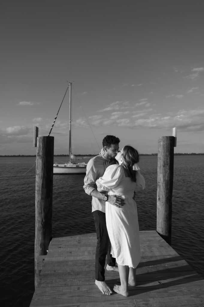 Black and white image of couple kissing on dock in Florida with their sailboat in the bay behind them