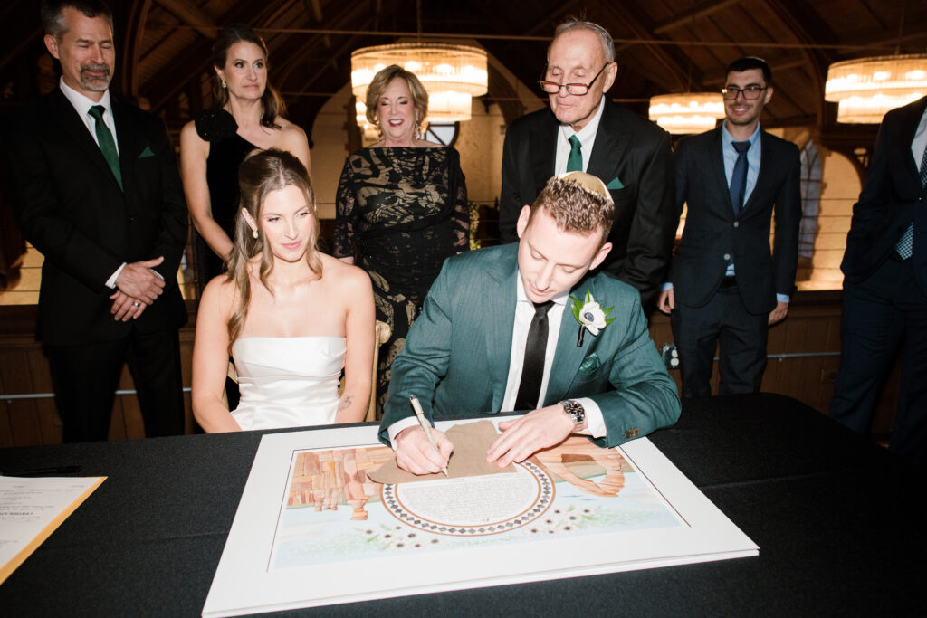 Bride and groom signing their Ketubah with their family behind them at The Palomar in Cincinnati, Ohio