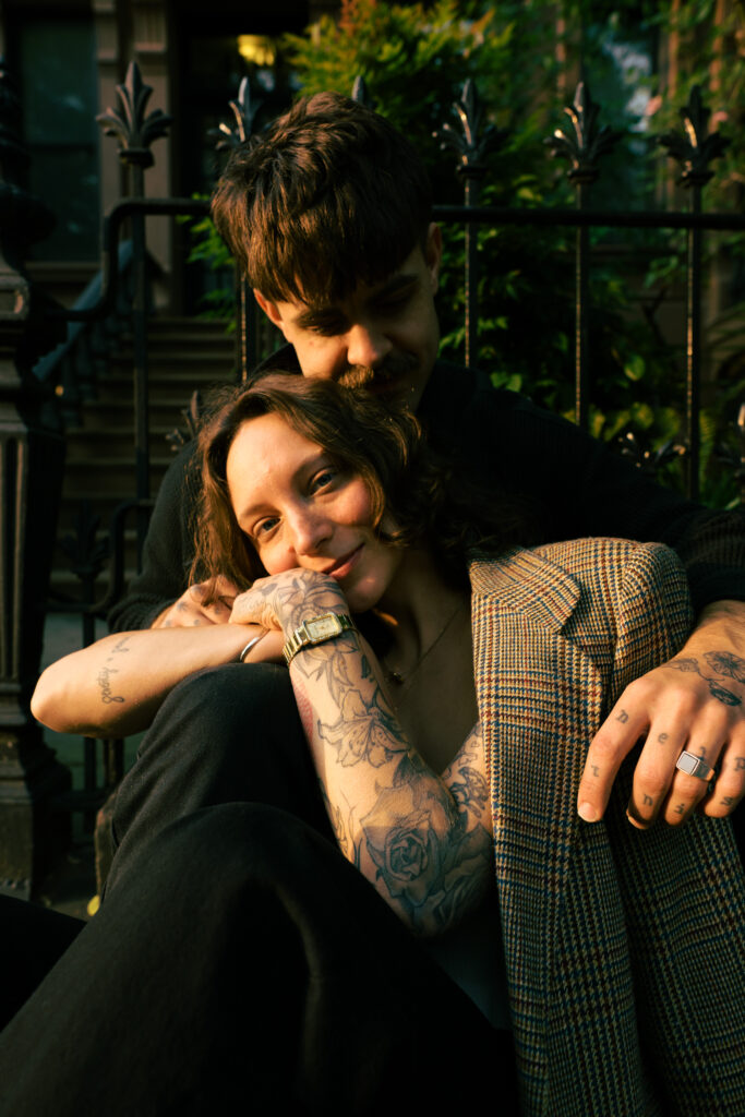 Bride snuggled up on grooms knee, looking directly into the camera with the sun on her face during her engagement session 