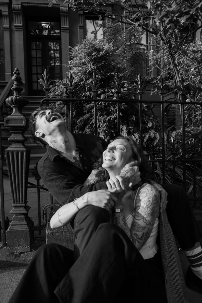 Black and white image of couple laughing while sitting on the sidewalk during their engagement session in Brooklyn, New York 