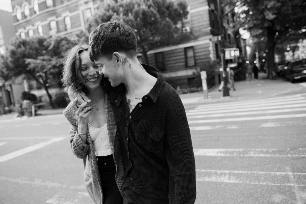 Black and white image of couple walking down the street with their arms around each other, laughing, during their Brooklyn  engagement session 