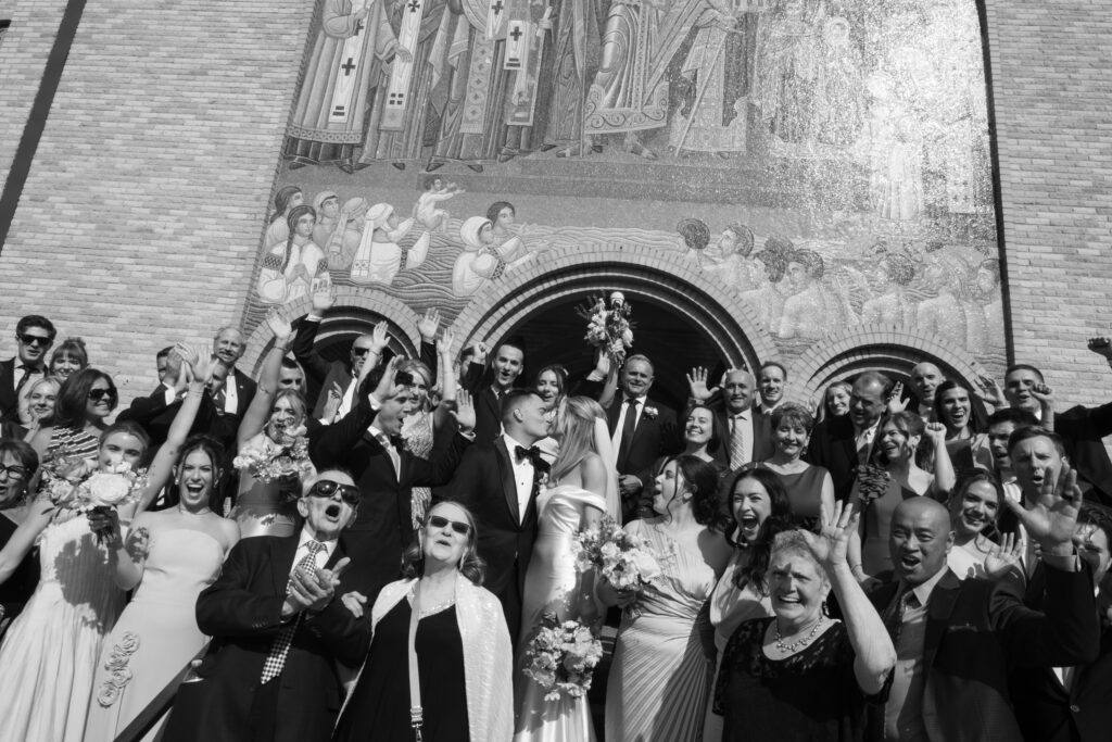 A black and white image of a couple kissing on the stairs of the church, right after their wedding ceremony, with all of their guests cheering around them 