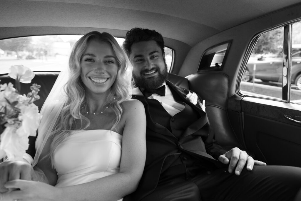 A black and white image of a couple, smiling in the back of their getaway car after their wedding ceremony 