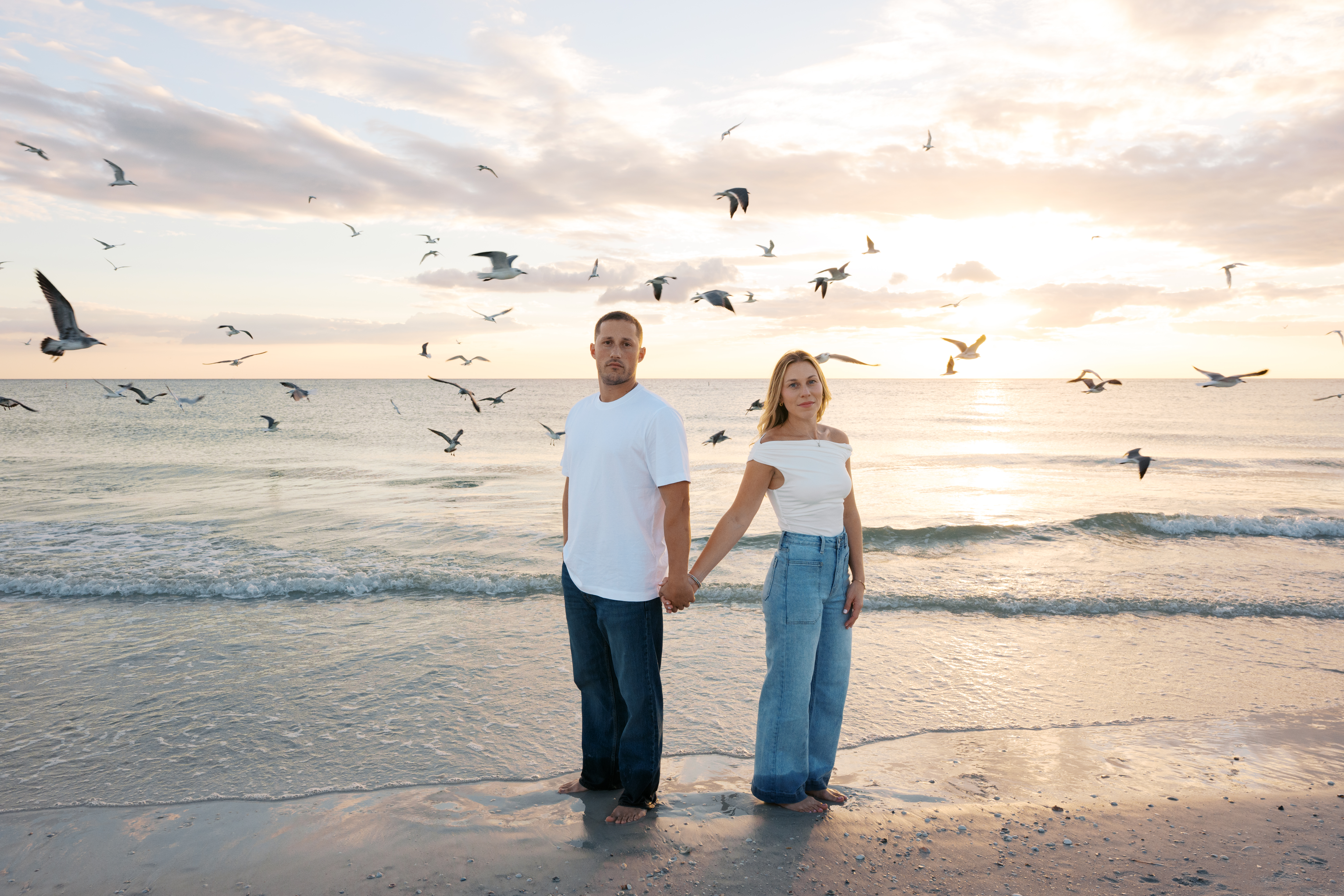 Couple holding hands and smiling at the camera during sunset on the beach with Seagulls flying behind them.