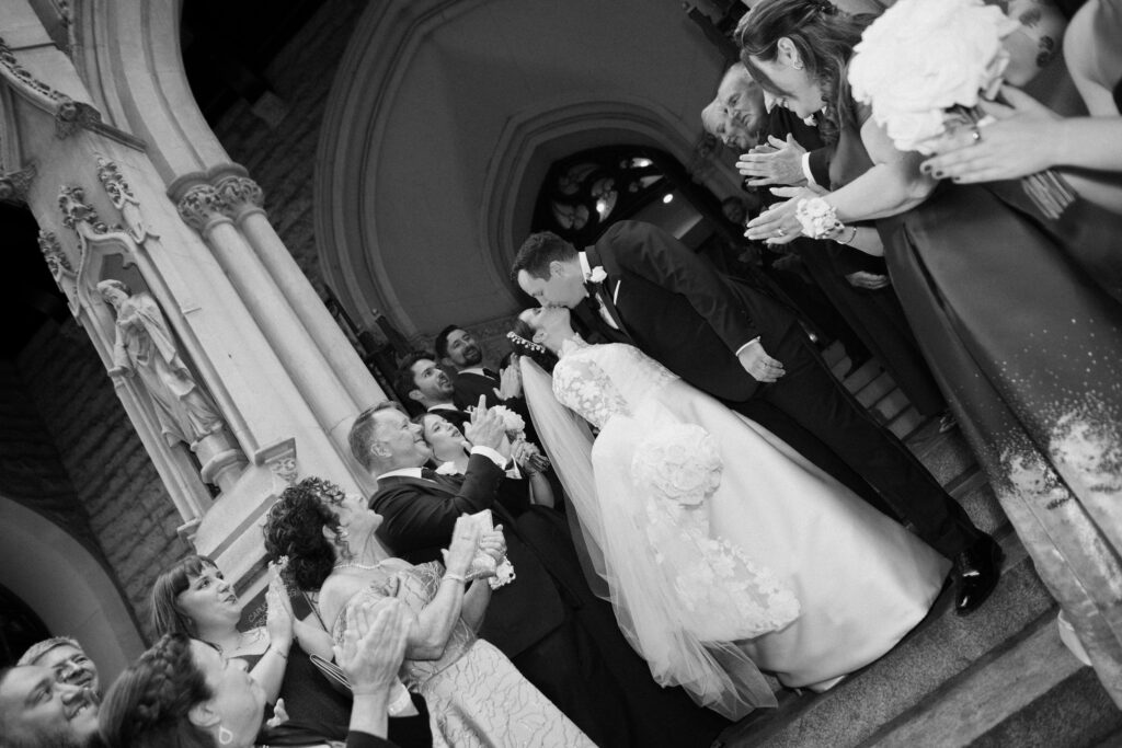 Black and white image of a couple kissing on the steps of the church with their family cheering around them captured by Wilmington Wedding Photographer Bri Nicole Photo Co.